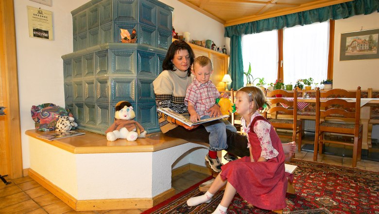 Relax by the fireplace, © Hausmann/Zwickl A woman sits with two children on a bench in front of a tiled stove and reads a book. Toys and decorations are scattered around her.