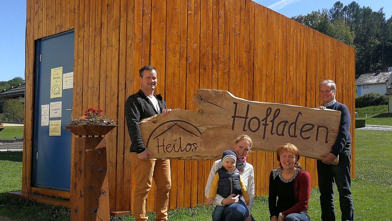 Ex farm sale Heilos-exterior view, © Claudia Gugler Family in front of a wooden building with a sign 'Heilos Hofladen'.
