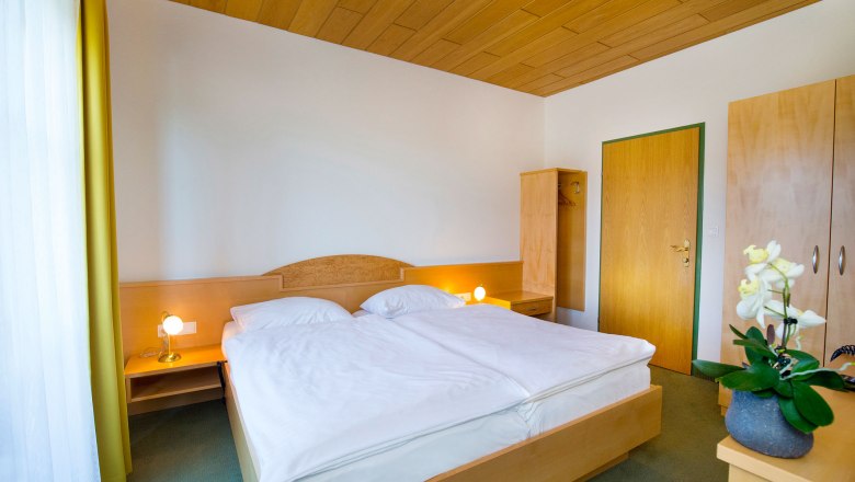 Room view, © Hotel Stich A simply furnished hotel room with a double bed, wooden ceiling, wardrobe and bedside lamps.