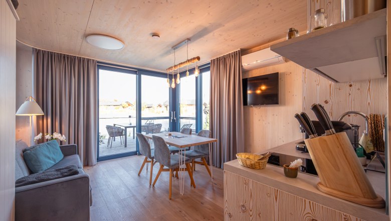 Kittenberger Chalets at the Gartensee, © Niederösterreich Werbung / Maximilian Pawlikowsky Interior view of a modern chalet with wooden walls, dining table, sofa and large window front overlooking a terrace.