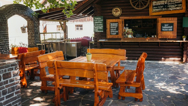 Barbecue evenings in the garden, © Niederösterreich Werbung/Daniela Führer Rustic guest garden with wooden tables and benches, surrounded by stone walls and a small bar area.