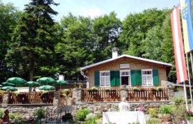 Falkensteinerhütte, © Falkensteinerhütte A mountain hut with green shutters, surrounded by trees, with a garden and parasols in the foreground.