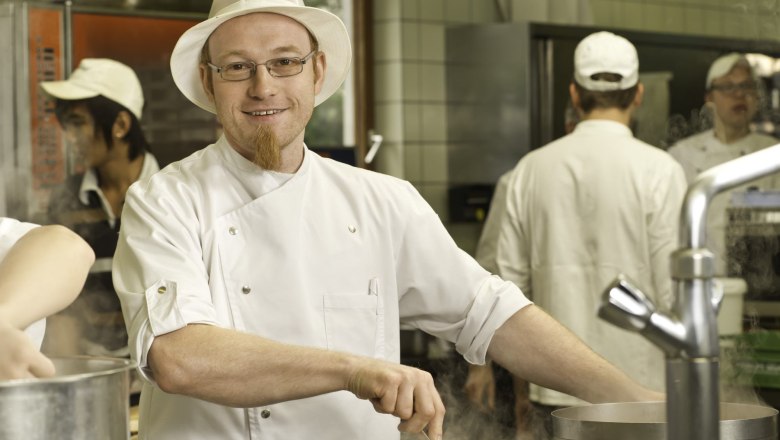 Josef Knoll, © Rita Newman A cook in a white uniform and hat stirs in a pot in a commercial kitchen.