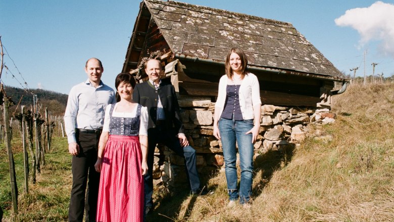 rusher-1, © Familie Rauscher Four people are standing in front of an old hut on a hill with vines in the background.