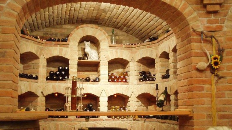 Rustic domed wine cellar, © Hans Krenn A wine cellar with brick shelves filled with bottles of wine. A cat figure stands in the middle. A sunflower hangs on the wall.