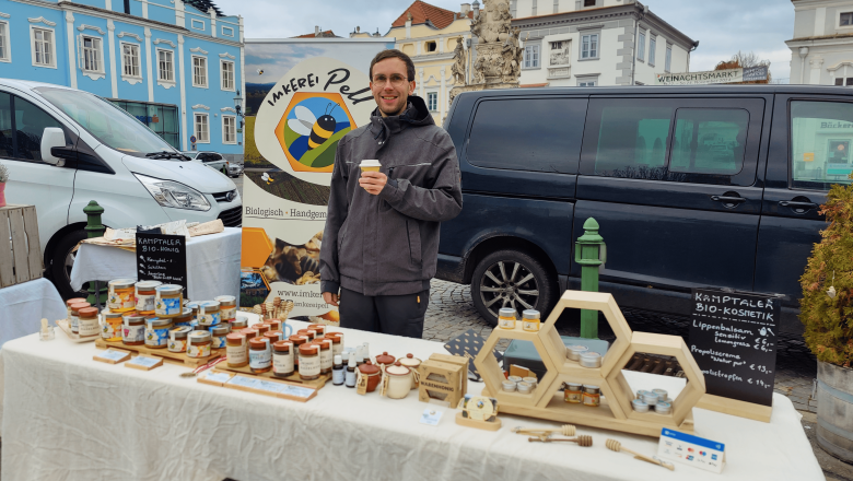 Alex at the market in Langenlois, © Imkerei Pell A man stands at a market stall with honey products and holds a cup. A van and buildings can be seen in the background.