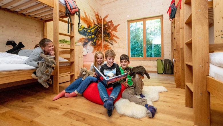 Wildcat Camp, © Waldviertel Tourismus / Studio Kerschbaum Children in a wooden room with bunk beds, reading a book and holding cuddly toys.