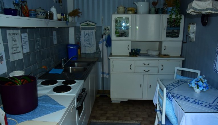 Supply, © Waldcamping Hubertus Small, cozy kitchen with blue and white decoration, a stove, sink, cupboard and small dining table.