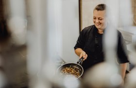 Takes care of the kitchen: Jessica Mann, © Niederösterreich Werbung/Sophie Menegaldo A cook in a black uniform holds a pan of roasted vegetables and smiles.