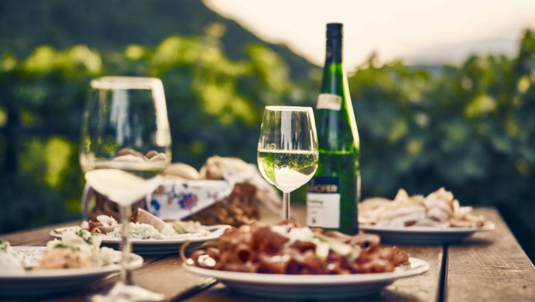 Heurigen snack, © Andreas Hofer A table with wine glasses, a bottle of wine and plates with various dishes outside.