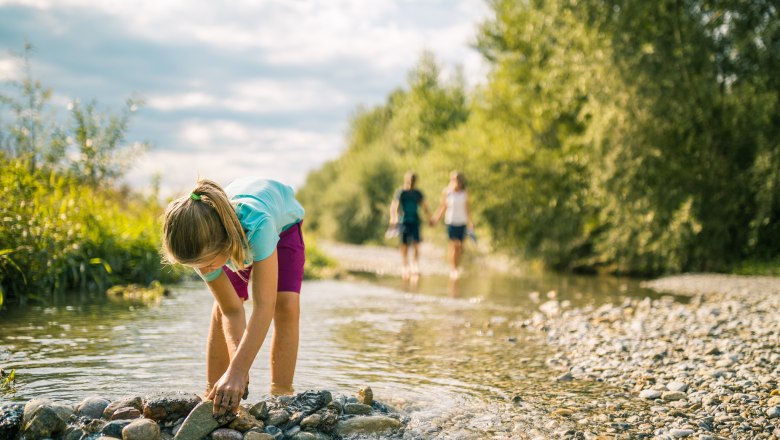 In the Leitha-Au, © Wiener Alpen, Martin Fülöp A child plays in a shallow stream while two people walk in the background.