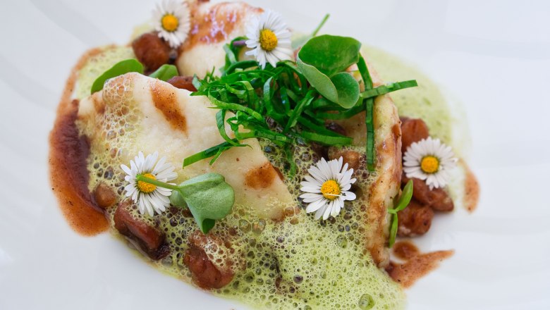 Delicacies from the award-winning kitchen, © Familie Görg Gourmet dish with green foam, daisies and herbs served on a white plate.