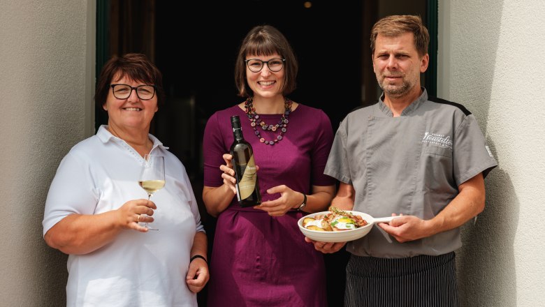 Wine.restaurant Neustifter, © Weinviertel Tourismus / Michael Reidinger Three people in front of a restaurant, a woman with a wine glass, a woman with a wine bottle, a man in a chef's outfit with a plate.