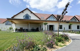 Woodland, © waldland A modern building with a red roof, surrounded by a well-tended garden with colorful flowers and lawn, under a blue sky.