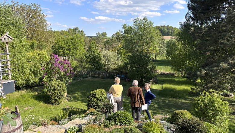 Gitti's garden paradise, © "Natur im Garten" Three people are standing in a green garden with trees and bushes, surrounded by a rural landscape under a blue sky with clouds.