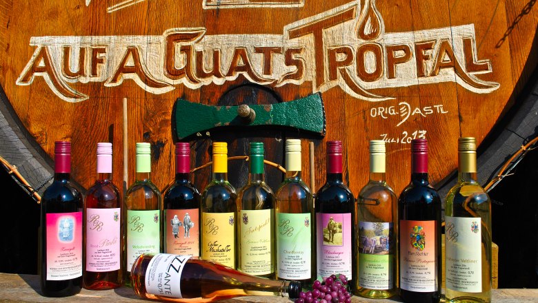 WineMood, © Heuriger Richter-Regenspurger Various bottles of wine in front of a large wooden barrel with the inscription 'Auf a guats Tropfal'.