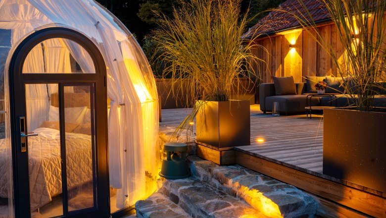 Chalets & places full of WONDER - Weidehaus De Luxe, © WUNDERVOLL eben A modern chalet with a terrace and a transparent dome tent in the forest at dusk.