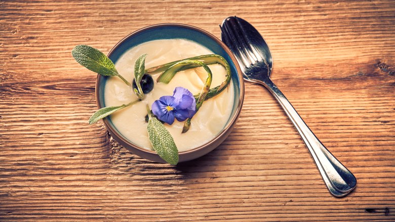 Cream of asparagus soup, © Niederösterreich Werbung/Andreas Hofer Cream of asparagus soup Soup with blossom and herbs on wooden table, spoon next to it.
