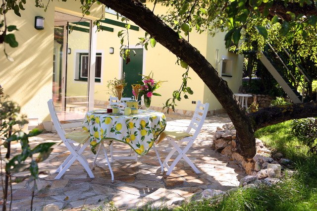 Terrace of the Romantik Cottage, © Frau Hahn A table with a lemon tablecloth and two white chairs on a terrace under a tree.