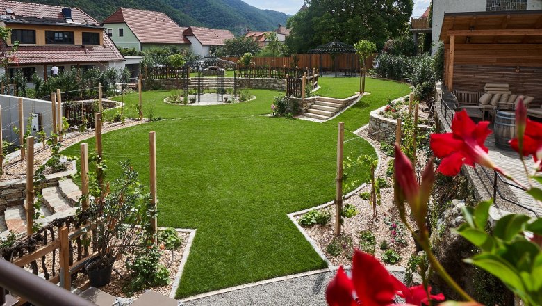 Kirchenwirt Wachau hotel garden, © Lehmann A well-tended hotel garden with a green lawn, flower beds and a pavilion, surrounded by houses and hills in the background.