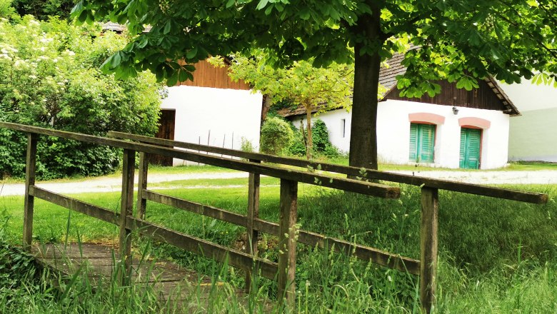 Romantic wine cellar lane in the middle of the Schmidatal valley, © Weinstraße Weinviertel A small wooden bridge leads across a meadow to white buildings with green doors, surrounded by trees and bushes.