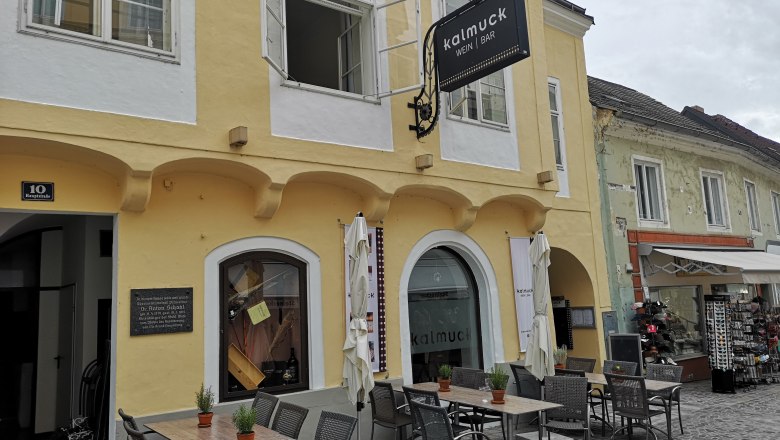 Exterior view of Kalmuck, © Donau NÖ Tourismus Exterior view of a yellow building with the sign 'Kalmuck Wine Bar'. There are tables and chairs in front of the building.