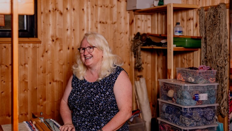 Arts and crafts Roswitha Haghofer, © Waldviertel Tourismus, Matthias Streibel A woman with blonde hair sits smiling in a room with wooden walls, surrounded by yarn and craft materials.