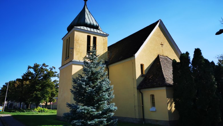 Hetzmannsdorf Chapel, © Weinstraße Weinviertel Yellow chapel with tower and cross, surrounded by trees, under a blue sky.