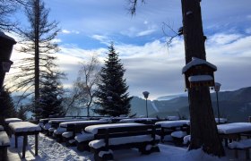 Terrace Speckbacher Huts, © Karin Stranz Snow-covered terrace with wooden benches and tables, surrounded by trees and mountains in the background.