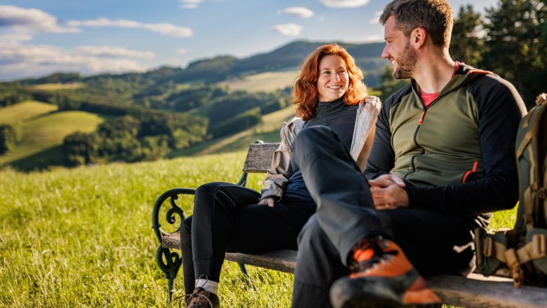 Am Bener in Klingfurth/Walpersbach, © Wiener Alpen/Fülöp, Kremsl A woman and a man in hiking gear are sitting on a wooden bench. Meadow, green hills and forest in the background.