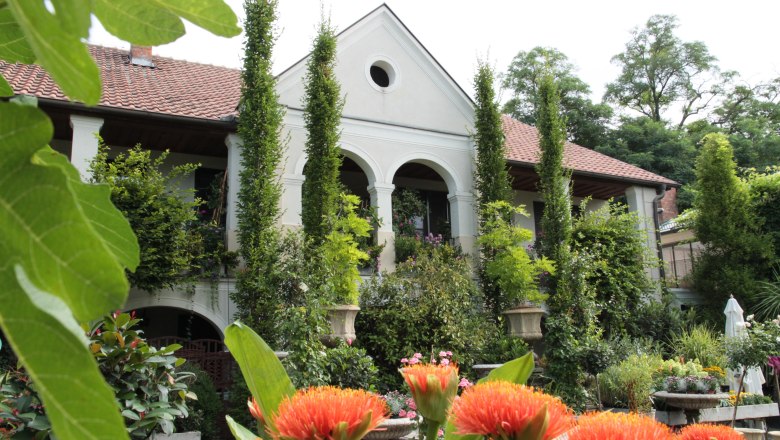 Nursery-Atzenbrugg, © Lederleitner A white building with a red tiled roof, surrounded by lush vegetation and orange flowers in the foreground.