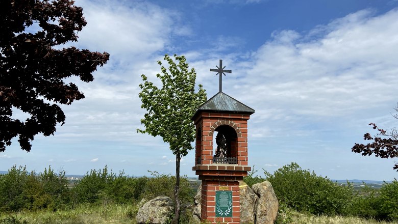 Lord's seat Etzmannsdorf, © Weinstraße Weinviertel A brick wayside shrine with a cross on a hill, surrounded by trees and bushes, under a blue sky.
