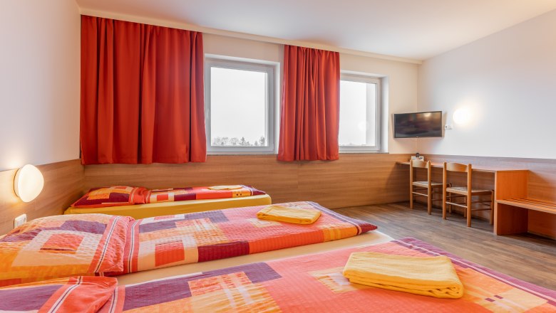 OEKOTEL Föhrenhain, © OEKOTEL Föhrenhain Hotel room with three beds, red curtains and a TV on the wall.