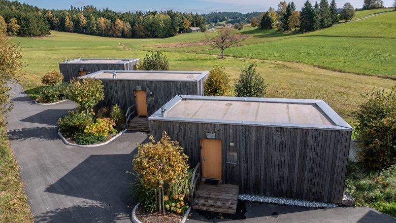roo'n, © Niederösterreich Werbung / Maximilian Pawlikowsky Three modern wooden houses in a green landscape with trees and meadows.