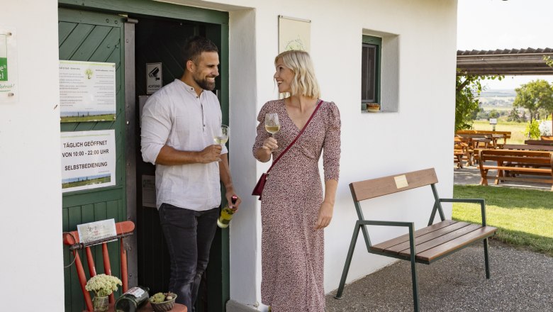 Self-service cellar at the Baumkreis, © Weinviertel Tourismus GmbH / Schwarz-König A man and a woman are standing in front of a small white building drinking wine.