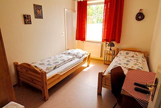 Room view, © Martin Herget A simply furnished room with two single beds, a window with red curtains and a small table and chair.