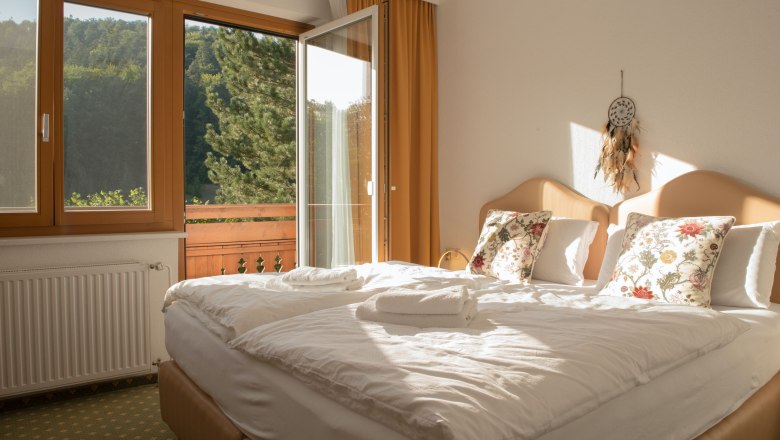 Single room with balcony, © Dorfschmiede eG A cozy single room with a double bed, decorative cushions and an open balcony with a view of green trees.
