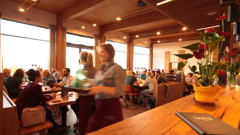 Guest room Kerbl am Weinberg, © Philipp Toscani A busy dining room with wooden furniture, guests at tables and a waitress in the foreground. A plant stands on the bar.