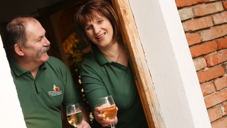 Tavern Angerbauer, © schwarz-koenig.at Two people in green polo shirts with wine glasses in their hands, smiling in a doorway.