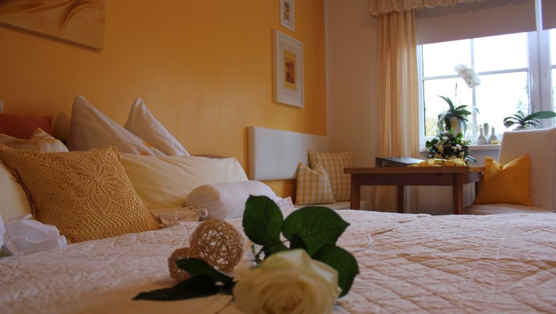Room, © Gästehaus Schabel-Zehetner Cozy bedroom with yellow walls, a bed with white and yellow pillows, a table and a window with plants.
