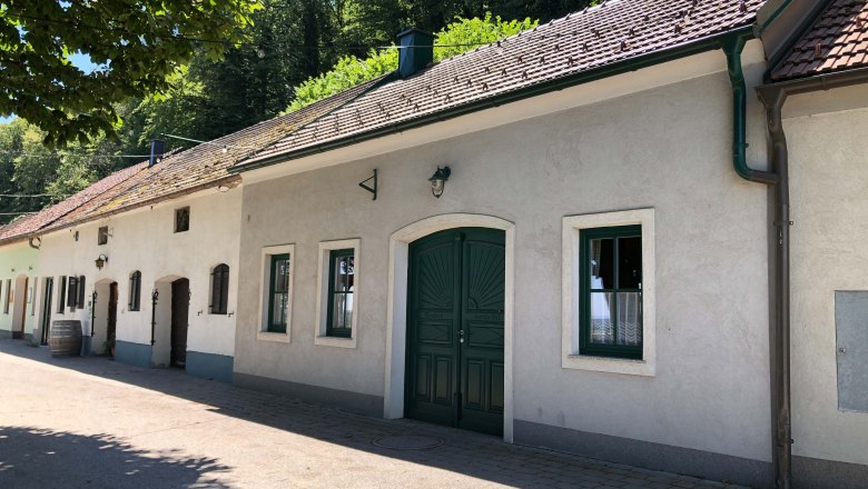Wine tavern outside, © Donau Niederösterreich Exterior view of a traditional wine tavern with white walls and green gate, surrounded by trees.