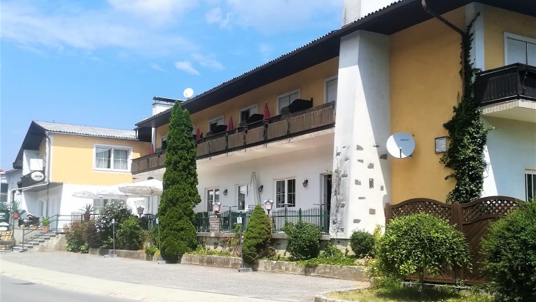 Bernhart pub stage, © Wiener Alpen A two-storey building with balcony and terrace, surrounded by plants and trees, in sunny weather.