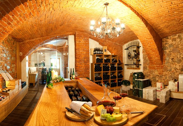 Wine cellar, © Residenz Wachau A cozy wine cellar with brick walls, a wooden table, shelves of wine bottles and a plate of cheese and grapes.