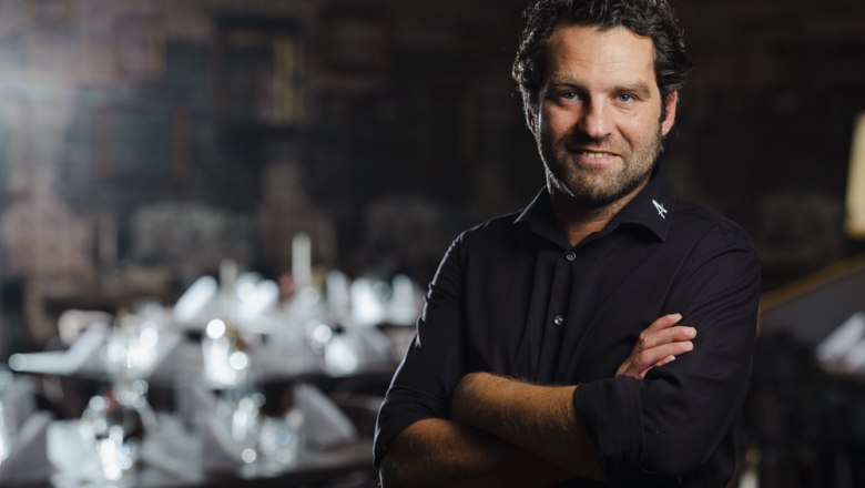 Show cooking courses with host Alexander Muhr, © Niederösterreich Werbung/Michael Reidinger Man in a black shirt stands in front of an elegantly laid table in a dark room.