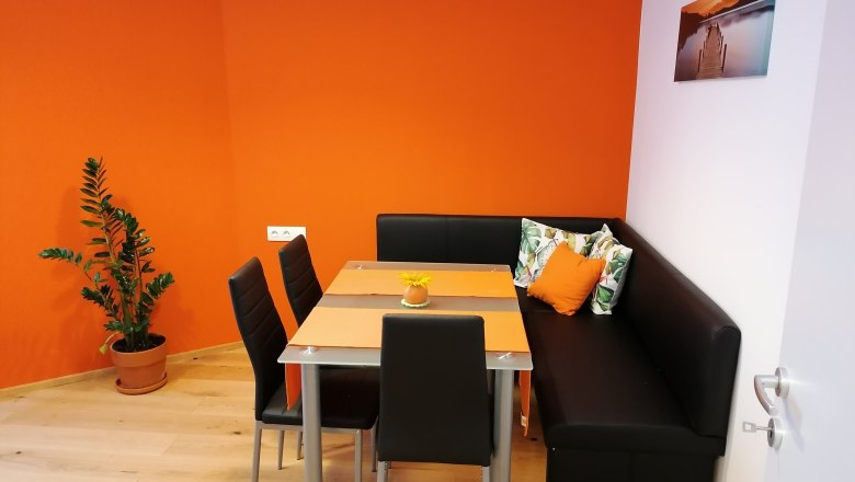 Breakfast room, © Pension zum Spitz A modern breakfast room with an orange wall, black corner bench, table and chairs. A plant stands in the corner, a picture hangs on the wall.