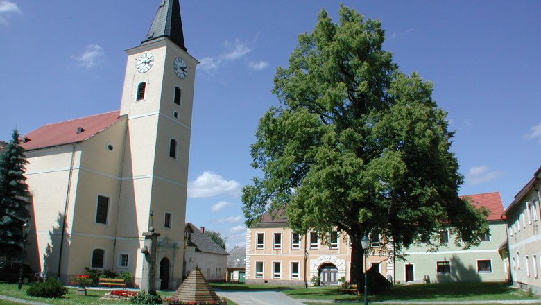 Hirschbach, © Hirschbach Church with tower and clock in a village square with trees and buildings.