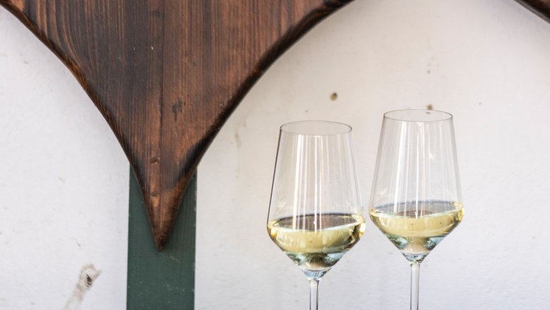 Wine glasses, © Weinviertel Tourismus / Doris Schwarz-König Two wine glasses with white wine on a wooden table in front of a wooden wall with heart decoration.