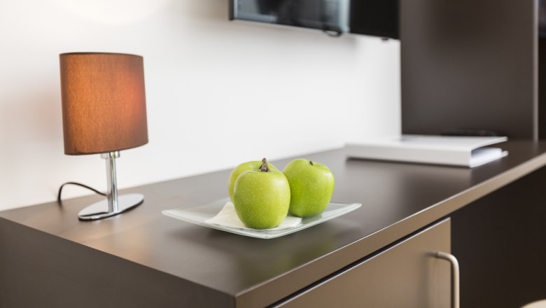 Guest room Jöchlinger, © Ernst Jöchlinger A table with three green apples on a plate, a lamp and a television in the background.