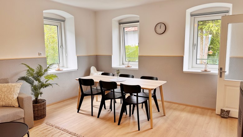 Living area, © Christoph Menhofer Dining area with wooden table and black chairs, light-colored walls, large windows, clock on the wall.