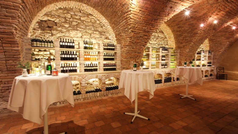 Vinotheque Mailberg, © Joseph Gasteiger A wine cellar with brick walls, shelves full of wine bottles and bar tables with white tablecloths.